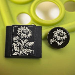 Könnte beinhalten: Zwei schwarze Dekorationsgegenstände mit einem weißen Sonnenblumenmuster. Eines ist rechteckig, das andere rund. Die Sonnenblume hat detaillierte Blütenblätter und Blätter. Der Hintergrund ist hellgrün.