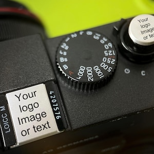 Könnte beinhalten: Nahaufnahme eines schwarzen Kameragehäuses mit einem silbernen Steuerungsrad und einem schwarzen Zifferblatt mit weißen Zahlen. Die Kamera hat eine silberne Platte mit dem Text "Your logo Image or text".