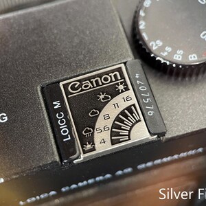 May include: A black camera top plate featuring a silver exposure guide with the Canon logo, weather icons, and aperture settings from 4 to 16. A black shutter speed dial with white numbers is visible, along with the text LOICC M and a serial number.