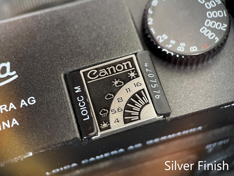May include: A black camera top plate featuring a silver exposure guide with the Canon logo, weather icons, and aperture settings from 4 to 16. A black shutter speed dial with white numbers is visible, along with the text LOICC M and a serial number.