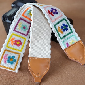 Könnte beinhalten: Ein weißer Kameragurt mit cremefarbenem Mittelteil und bunten, gestickten Blumenquadraten. Der Gurt hat braune Lederenden und einen braunen Riemen zur Befestigung. Die gestickten Blumen sind in Rosa-, Orange-, Gelb-, Blau- und Lilatönen gehalten.