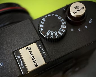 May include: Close-up of a black Fujifilm camera body. The camera features a silver dial with numbers and a silver Fujifilm logo. The top of the camera has a silver button with the Fujifilm logo and the text "FUJIFILM".