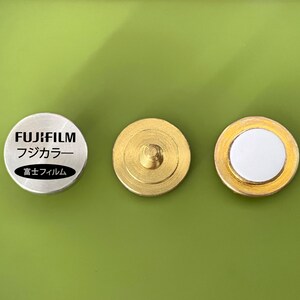 May include: Three round metal objects. The left object is silver with the text "FUJIFILM" and "フジカラー" in black. The right two objects are gold with a white center.