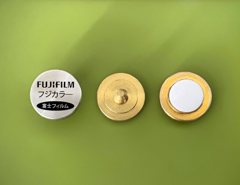May include: Three round metal objects. The left object is silver with the text "FUJIFILM" and "フジカラー" in black. The right two objects are gold with a white center.