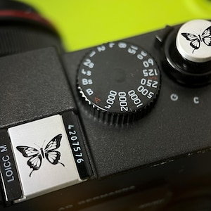 May include: Close-up of a black camera body with a silver butterfly emblem. The camera features a black dial with white numbers and a silver button with a butterfly design. The text "LOICC M" and "4207576" are visible.