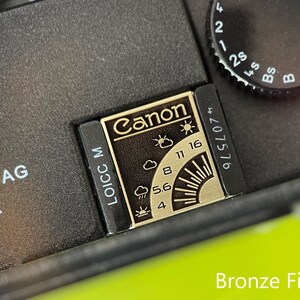 May include: A black camera accessory with a bronze finish metal plate featuring the Canon logo, weather icons, and aperture settings including 4, 5.6, 8, 11, and 16. The plate is marked with LOICC M and a serial number, positioned next to a shutter speed dial.