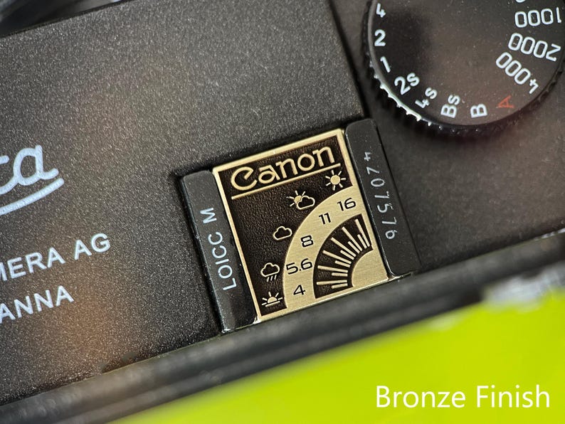 May include: A black camera accessory with a bronze finish metal plate featuring the Canon logo, weather icons, and aperture settings including 4, 5.6, 8, 11, and 16. The plate is marked with LOICC M and a serial number, positioned next to a shutter speed dial.