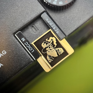 May include: Close-up of a black camera with a gold-colored accessory. The accessory features a detailed illustration of a skeleton wearing a hat. The camera has the text "Leica Camera AG Wetzlar, Germany" and the number "407576".