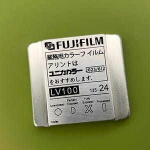 May include: A silver metal film canister with black text that reads "FUJIFILM", "業務用カラーフイルム", "アリントは", "ユニカカラー", "2023/6/2", "をおすすめします.", "LV100", "135-24", "Partially", "Fully", "Unexposed", "Exposed", "Exposed", "Processed", "Noles".