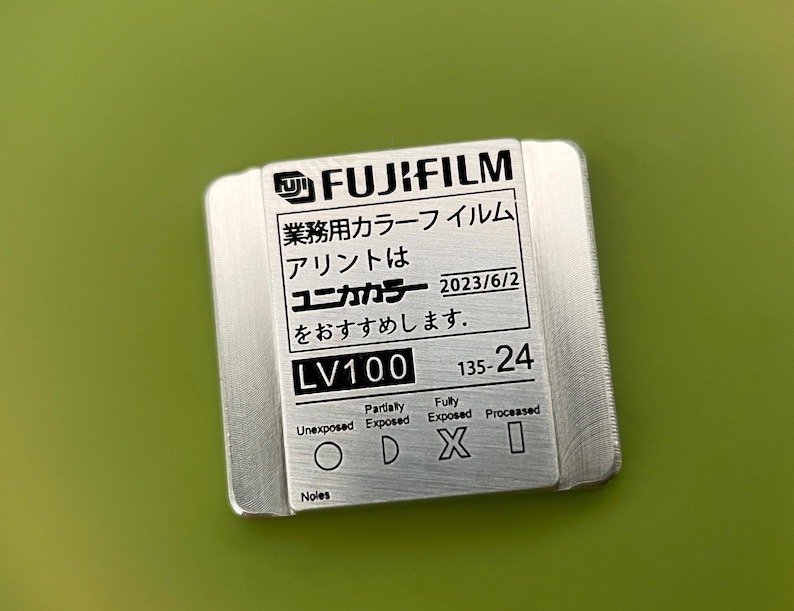 May include: A silver metal film canister with black text that reads "FUJIFILM", "業務用カラーフイルム", "アリントは", "ユニカカラー", "2023/6/2", "をおすすめします.", "LV100", "135-24", "Partially", "Fully", "Unexposed", "Exposed", "Exposed", "Processed", "Noles".