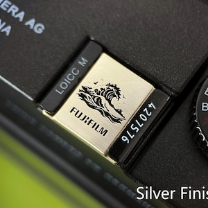 Op de afbeelding: Close-up van een zilveren Fujifilm cameradetail. De zilveren plaat heeft een golfontwerp en het Fujifilm-logo. Zwarte tekst bevat "LOICC M" en "4207576". De camerabody is zwart, met een draaiknop en de tekst "Silver Finish".