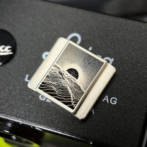 Op de afbeelding: Zilverkleurige hot shoe cover voor camera's met een berg- en zonontwerp. Het gedetailleerde landschap is gegraveerd met fijne lijnen. De vierkante cover heeft afgeronde hoeken en ligt op een zwarte camerabody.