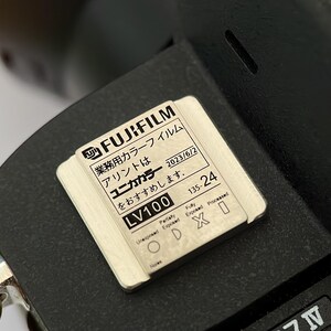 May include: A close-up of a black camera with a white label that reads "FUJIFILM 業務用カラーフイルム アリントは ユニカカラー をおすすめします。 LV100 135-24 2023/6/2".