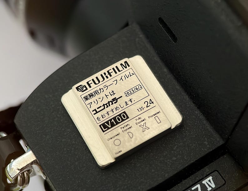 May include: A close-up of a black camera with a white label that reads "FUJIFILM 業務用カラーフイルム アリントは ユニカカラー をおすすめします。 LV100 135-24 2023/6/2".