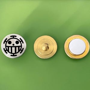 May include: Three round metal buttons. The first button is silver with a black and white skull and crossbones design. The second button is gold with a small metal post in the center. The third button is gold with a white center.