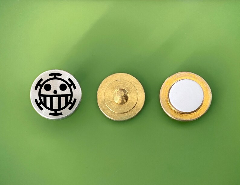 May include: Three round metal buttons. The first button is silver with a black and white skull and crossbones design. The second button is gold with a small metal post in the center. The third button is gold with a white center.