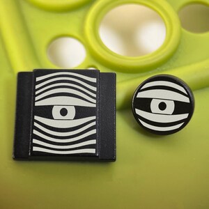 May include: A black square button and a black circular button, both featuring a white graphic design of a stylized eye surrounded by horizontal wavy lines. The buttons have a matte finish and are displayed together as a set of two distinct shapes.