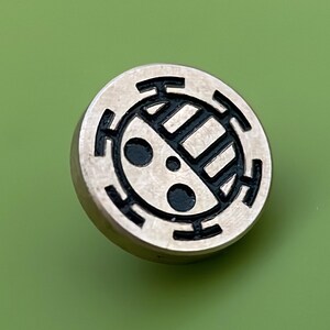 May include: A silver-colored metal pin with a black and white design. The design features a circle with a series of concentric rings and a stylized symbol inside.