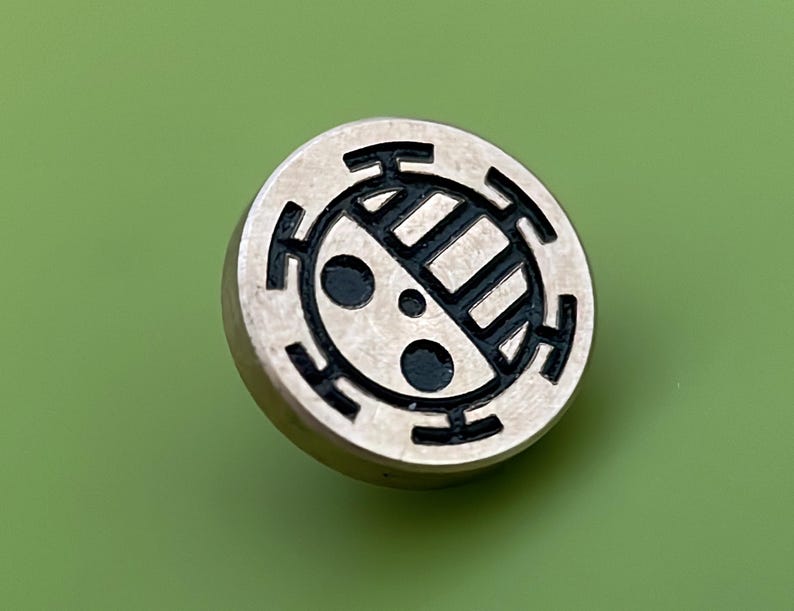 May include: A silver-colored metal pin with a black and white design. The design features a circle with a series of concentric rings and a stylized symbol inside.