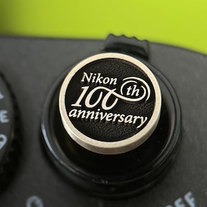 May include: A close-up shot of a black camera dial featuring a silver-edged emblem. The emblem displays "Nikon 100th anniversary" in white text. The dial is set against a blurred green backdrop.