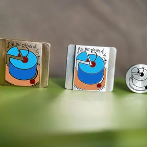 Op de afbeelding: Twee rechthoekige metalen pins en één ronde metalen pin met een illustratie van een blauwe taart met een kers erop en de Franse tekst J'ai des rêves à vivre. De rechthoekige pins hebben een goud- en zilverkleurige afwerking, de ronde is zilver.