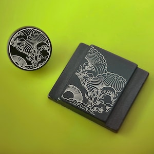 May include: A black round object and a black square object with a white wave and floral design. The objects are on a bright green surface. The round object is a plug. The square object is a coaster.