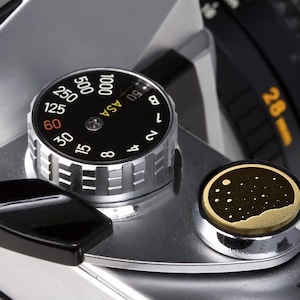 May include: Close-up of a silver camera with a black dial that reads "ASA" and has numbers from 6 to 1000. The dial is set to 60. There is a black and gold button with a starry night design.