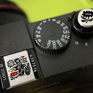 Könnte beinhalten: Eine schwarze Kameraoberseite mit einem runden Verschlusszeiten-Einstellrad mit weißen Zahlen, einem silbernen Auslöser mit schwarzem Blumensymbol und einer rechteckigen Metallplatte mit der Aufschrift LOICC M, der Nummer 4207576 sowie einer detaillierten schwarz-weiß-roten Illustration von Sonne, Blumen und abstrakten Formen.