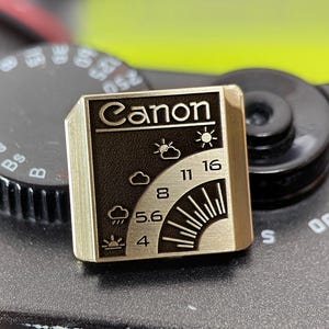 May include: A square metal pin featuring a gold-toned finish with a black recessed area. The design displays the word Canon at the top, followed by weather icons and aperture numbers 4, 5.6, 8, 11, and 16 arranged in a curved pattern.