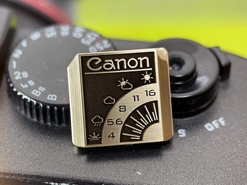May include: A square metal pin featuring a gold-toned finish with a black recessed area. The design displays the word Canon at the top, followed by weather icons and aperture numbers 4, 5.6, 8, 11, and 16 arranged in a curved pattern.