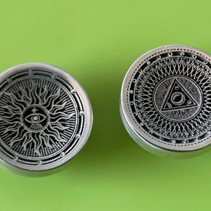 Op de afbeelding: Twee ronde knoppen van zilver met ingewikkelde ontwerpen. De knop links heeft een zon met een oog in het midden. De knop rechts heeft een driehoek met een oog in het midden, omringd door een cirkelvormig patroon.