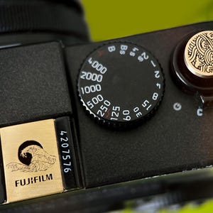 May include: Close-up of a black camera with a gold-colored dial and a gold-colored plate with the Fujifilm logo and a wave design. The camera has several dials with numbers and settings.