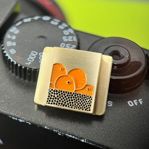 May include: A gold-coloured square pin with a design of orange fruit above a black dotted pattern. The pin is set against a black camera body with a dial and a brown knob.