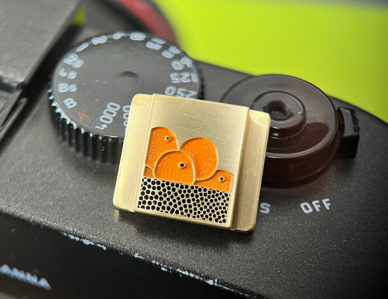 May include: A gold-coloured square pin with a design of orange fruit above a black dotted pattern. The pin is set against a black camera body with a dial and a brown knob.