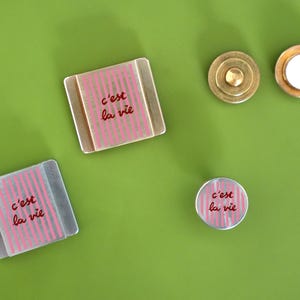 K&ouml;nnte beinhalten: Eine Sammlung dekorativer Magnete und ein Messingknopf auf einer gr&uuml;nen Oberfl&auml;che. Die Magnete haben ein rosa gestreiftes Design mit dem Schriftzug "c'est la vie" in Rot. Ein Magnet ist rund, zwei sind quadratisch.