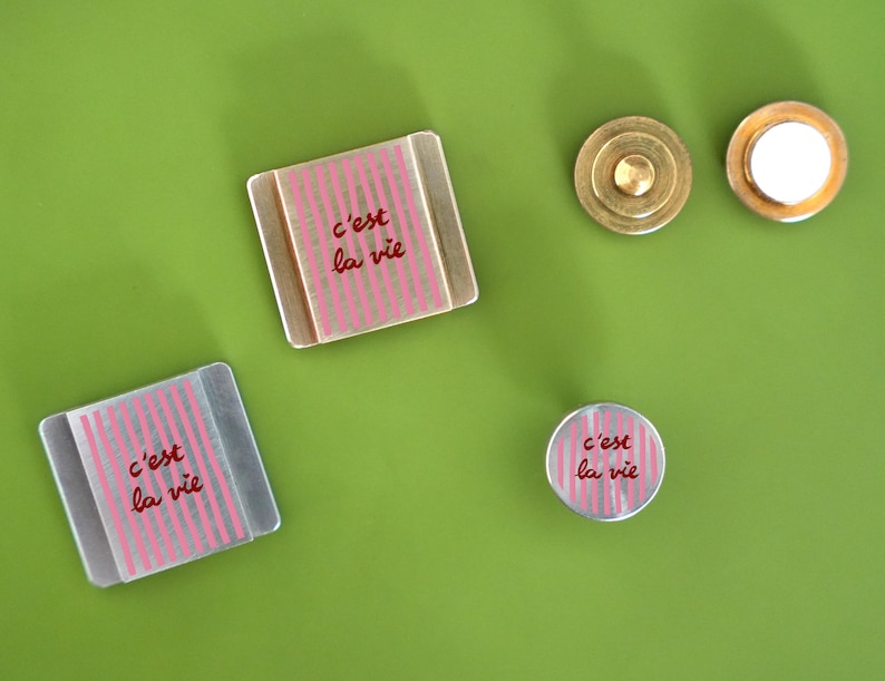 K&ouml;nnte beinhalten: Eine Sammlung dekorativer Magnete und ein Messingknopf auf einer gr&uuml;nen Oberfl&auml;che. Die Magnete haben ein rosa gestreiftes Design mit dem Schriftzug "c'est la vie" in Rot. Ein Magnet ist rund, zwei sind quadratisch.