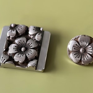May include: Two silver metal flower-shaped buttons with a detailed design. The larger button is rectangular and has multiple flowers, while the smaller button is round and features a single flower.