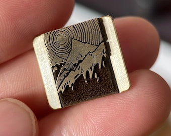 May include: A small, square metal object with a mountain and sun design. The mountains are etched in a darker color, while the sun and border are a lighter, metallic hue. The object is held between fingers.