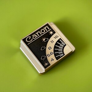May include: A square metal pin featuring the Canon logo in white text on a black background. The design includes a weather-themed exposure guide with icons for sun, clouds, and rain, alongside aperture numbers 4, 5.6, 8, 11, and 16 in silver.
