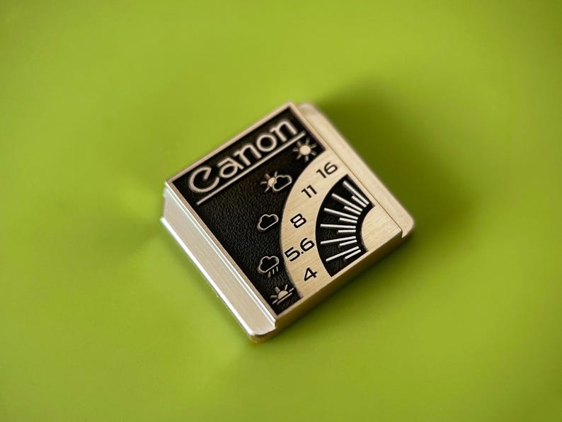 May include: A square metal pin featuring the Canon logo in white text on a black background. The design includes a weather-themed exposure guide with icons for sun, clouds, and rain, alongside aperture numbers 4, 5.6, 8, 11, and 16 in silver.