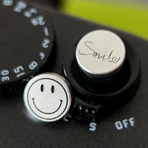 May include: Close-up of a black camera with silver dials and buttons. One button has a smiley face, and another has the word "Smile" in cursive. The camera has settings for different exposure times.