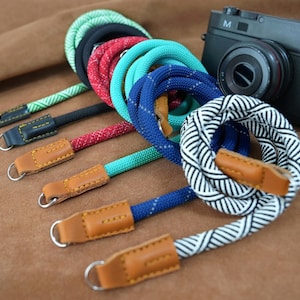 May include: A selection of camera straps in various colours, including green, black, red, turquoise, blue, and a black and white pattern. Each strap has a leather connector and a metal ring. A black camera is visible.
