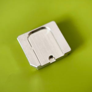 May include: A square, silver-colored metal plate with a brushed finish. The object features a recessed, U-shaped central area and a small, semi-circular cutout along one of its edges, showcasing a clean, industrial design with beveled corners.
