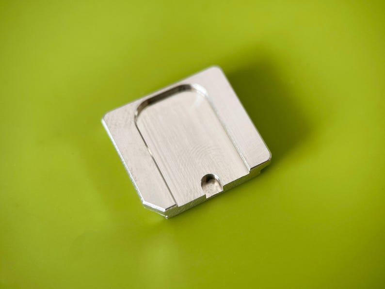 May include: A square, silver-colored metal plate with a brushed finish. The object features a recessed, U-shaped central area and a small, semi-circular cutout along one of its edges, showcasing a clean, industrial design with beveled corners.