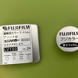 May include: A silver metal film canister with the text "FUJIFILM" and "LV100 135-24" printed on it. The canister has a black and white diagram showing the different stages of film exposure.  The text "Unika Color" is also printed on the canister.