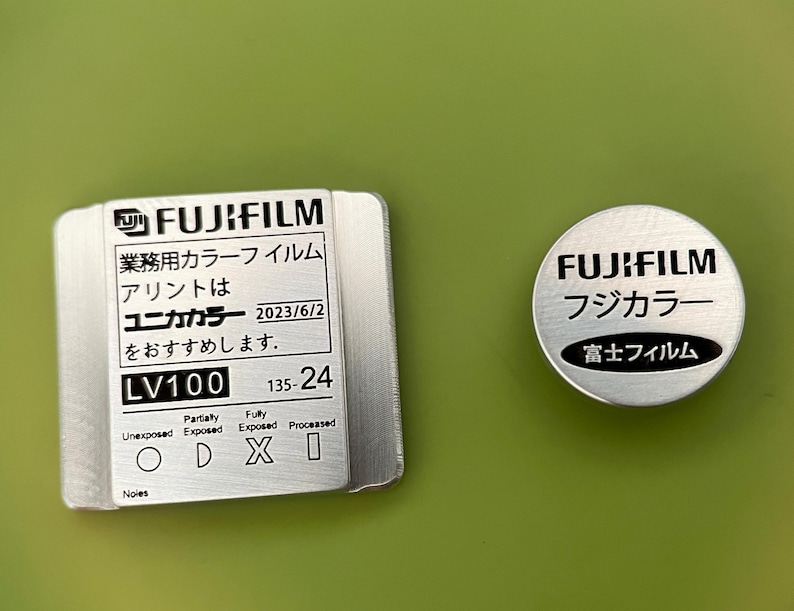 May include: A silver metal film canister with the text "FUJIFILM" and "LV100 135-24" printed on it. The canister has a black and white diagram showing the different stages of film exposure.  The text "Unika Color" is also printed on the canister.