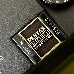 Könnte beinhalten: Nahaufnahme eines schwarzen Kameragehäuses mit einem metallischen Emblem. Das Emblem zeigt das Wort "PENTAX" über "100 YEARS OF HISTORY" in weißer Schrift. Eine Seriennummer und anderer Text sind ebenfalls sichtbar.
