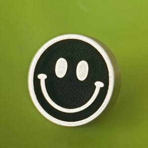 Puede incluir: Un pin redondo plateado con un centro negro que presenta una carita sonriente blanca. El pin tiene un borde metálico en relieve y está sobre un fondo verde liso. El diseño es simple y alegre.