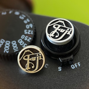 May include: Close-up of a camera with two silver and black Fuji logo buttons and a black dial with white numbers. The buttons are round with the Fuji logo and mountain design. The camera body is black.