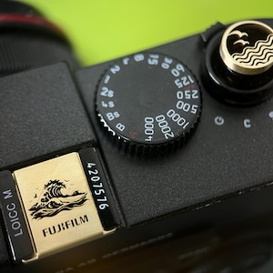 Op de afbeelding: Close-up van een zwarte camera met een goudkleurig accent met een golf- en vogelontwerp. De camera heeft een zwarte draaiknop met witte cijfers en een goudkleurige plaat met het woord "FUJIFILM".